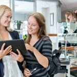 Beauty business owners looking over monthly numbers on a tablet while a hairdresser works in the background.