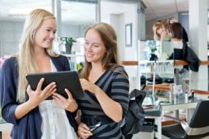 Beauty business owners looking over monthly numbers on a tablet while a hairdresser works in the background.